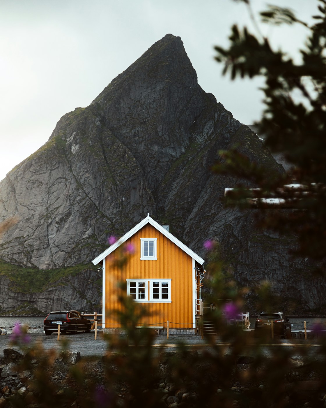 A small golden timber cabin on the Norwegian coast beneath a steep mountain.